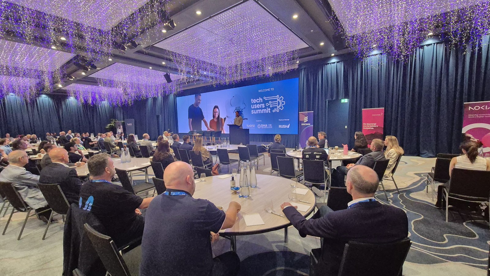 The Grand Millennium ballroom before the Tech Users Summit begins, delegates seated at round tables facing a stage screen reading Welcome to Tech Users Summit