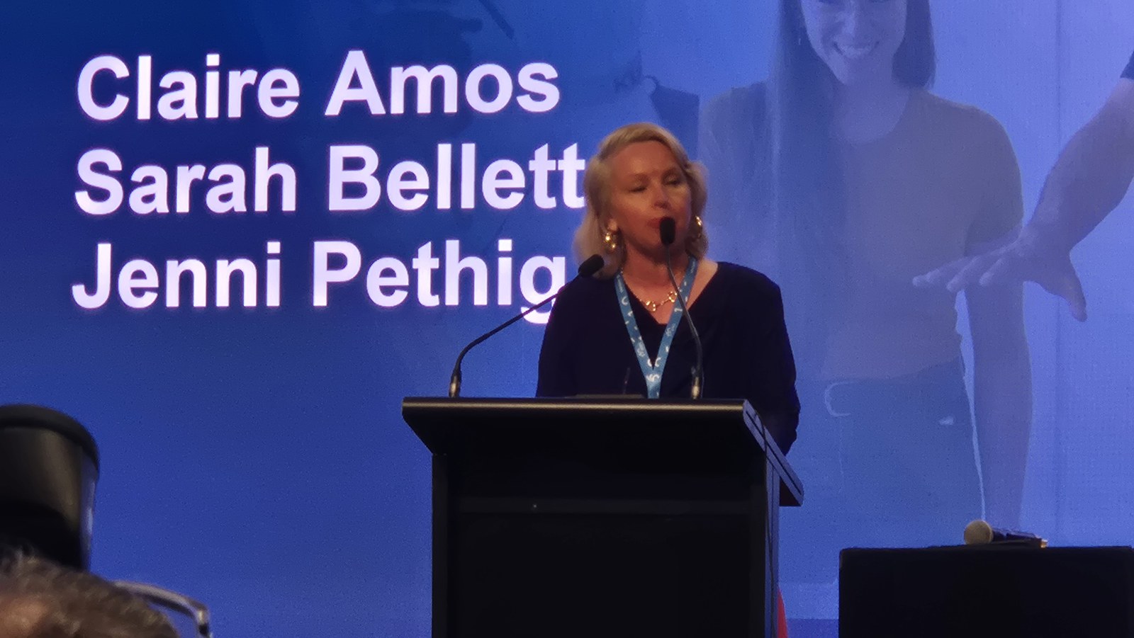 Claire Amos speaking at the lectern with the session title slide behind her listing Claire Amos, Sarah Bellett and Jenni Pethig